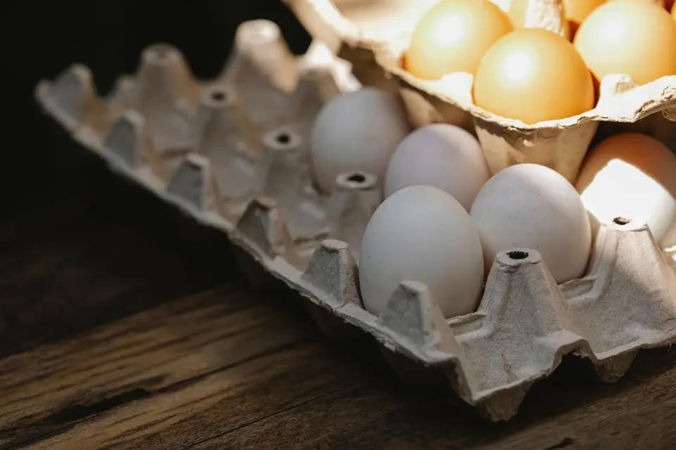 A pressed-paper egg carton holding a dozen brown eggs on a plain wooden table, lit by soft daylight.
