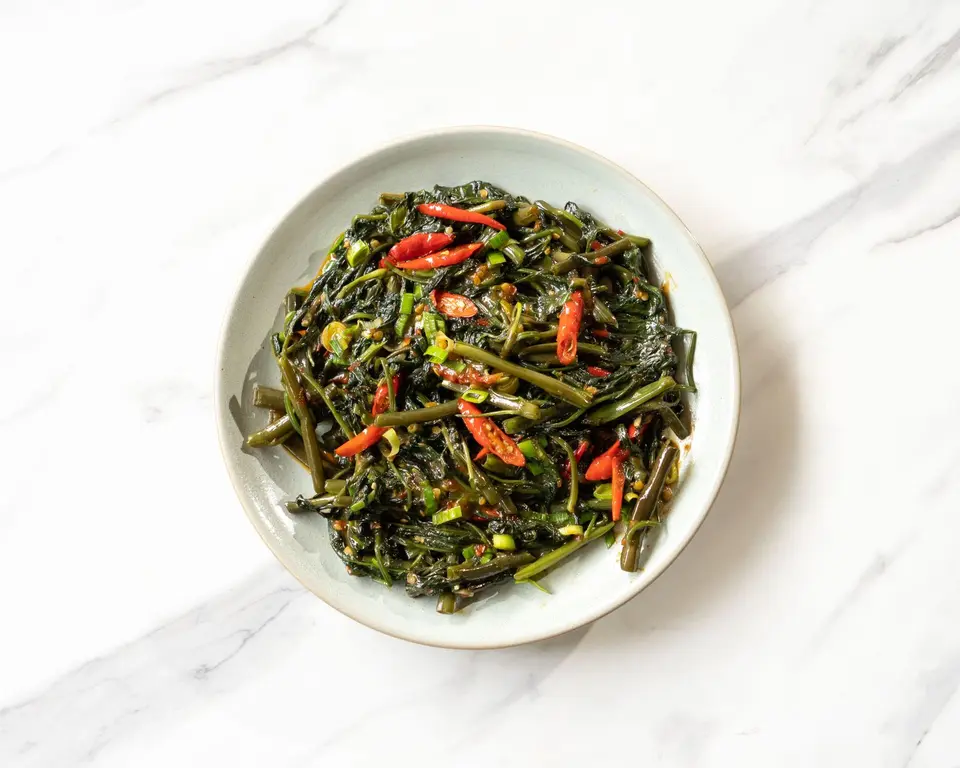 A plate of glossy stir-fried morning glory with whole garlic cloves and red chilli slices.