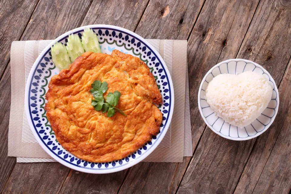 A golden puffed Thai omelette on a plate of steamed rice with a small bowl of chilli sauce.