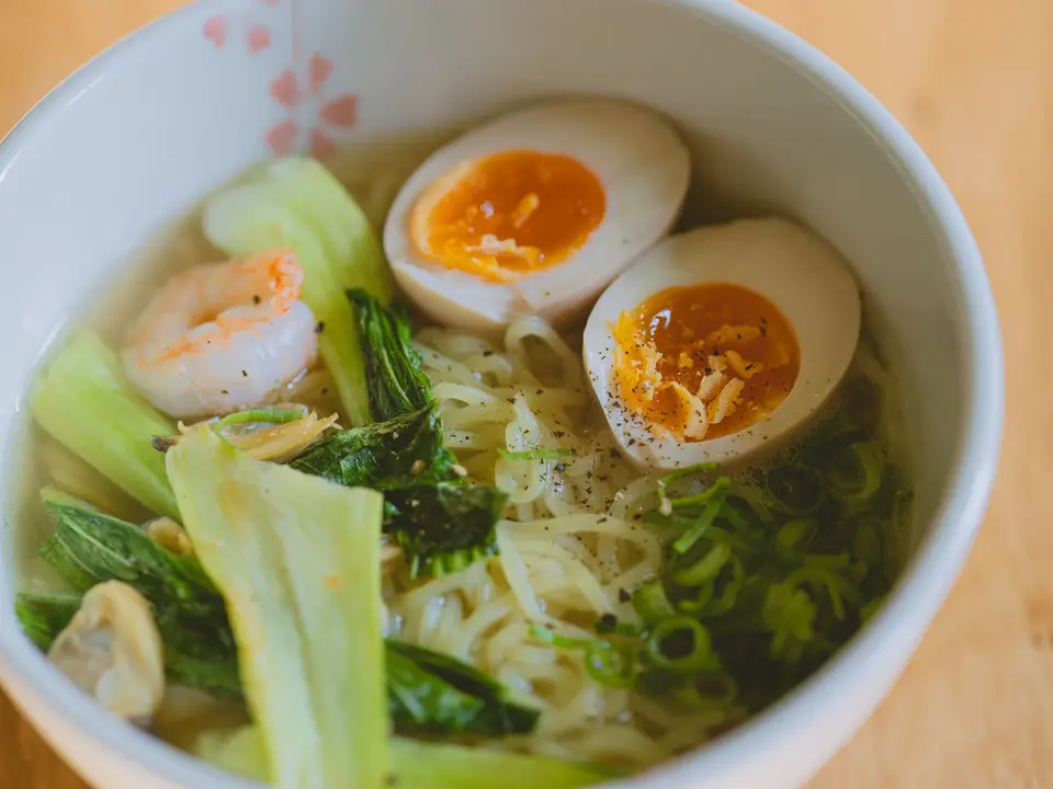 A white bowl of noodle soup with bok choy, prawns, sliced spring onion and a halved boiled egg.